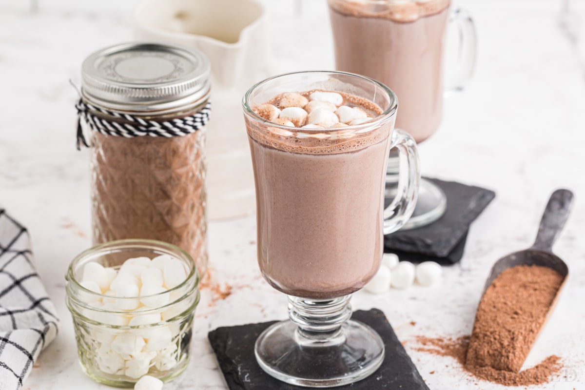 Two glasses of hot cocoa topped with mini marshmallows, glass jar with homemade hot cocoa mix with decorative ribbons, glass jar with mini marshmallows, checkered linen, metal cup with hot cocoa mix, on a marble countertop.