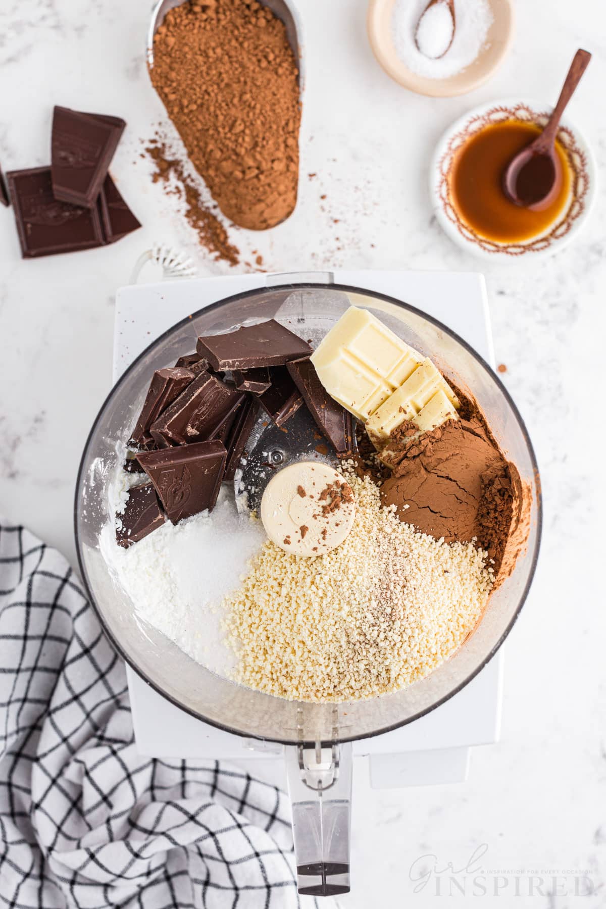 Hot cocoa ingredients added to a food processor, checkered linen, pieces of semi-sweet chocolate bar, metal measuring cup with unsweetened cocoa powder, bowl of sea salt, on a marble countertop.