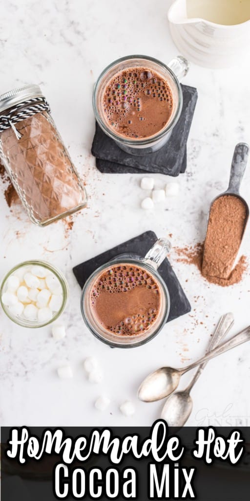 overhead shot of two glass mugs full of hot cocoa made with homemade hot cocoa mix, a milk jug, a glass jar of marshmellows, measuring spoon containing hot cocoa mix and two spoons on marble counter top