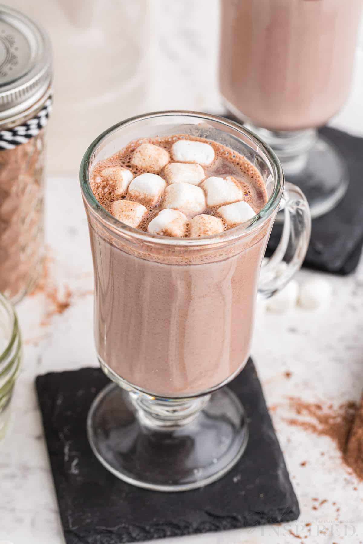 Two tall glasses with hot cocoa and mini marshmallow topping on top of black coasters, glass sealed jar filled with homemade hot cocoa mix, on a marble countertop.