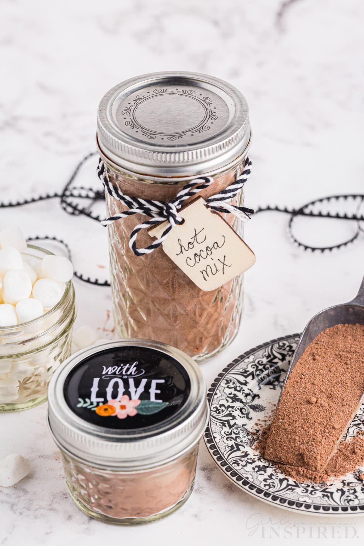 Two glass jars with homemade hot cocoa mix, decorative ribbon, and gift tags, glass jar with mini marshmallows, measuring cup with homemade hot cocoa mix, on a marble countertop.