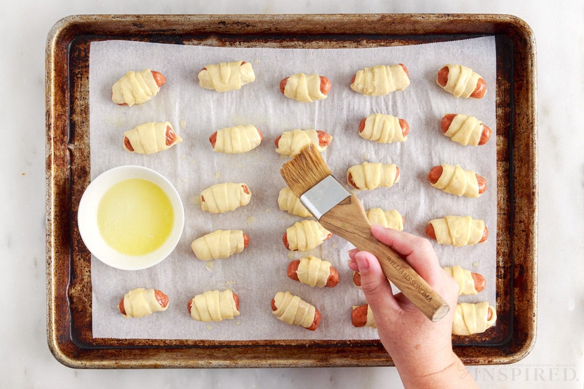 small dish of butter being brushed on top of lil smokies pigs in a blanket on a parchment paper lined cookie sheet