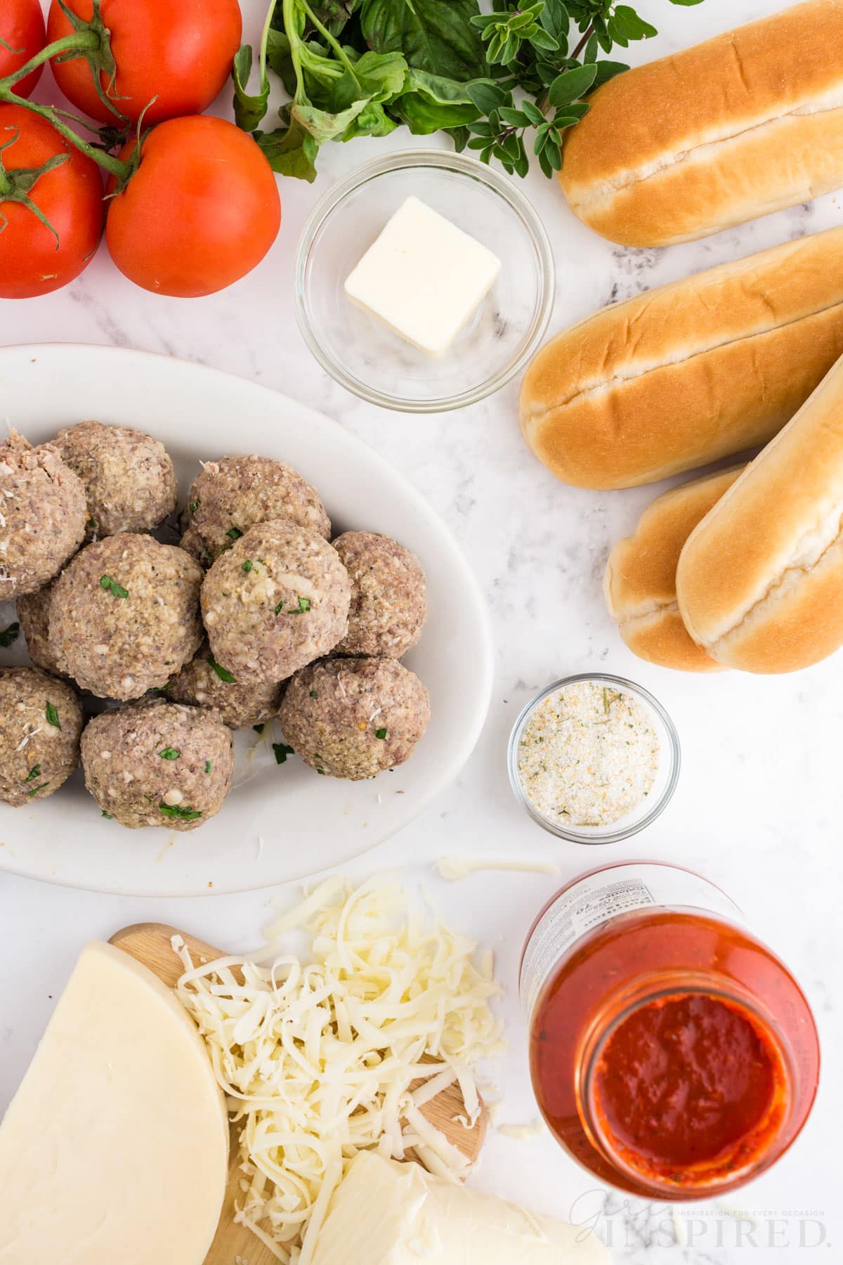 Plate of meatballs, wooden kitchen board with shredded cheese, marinara sauce, bread buns, tomatoes, basil, bowl of butter, garlic powder, on a marble countertop.