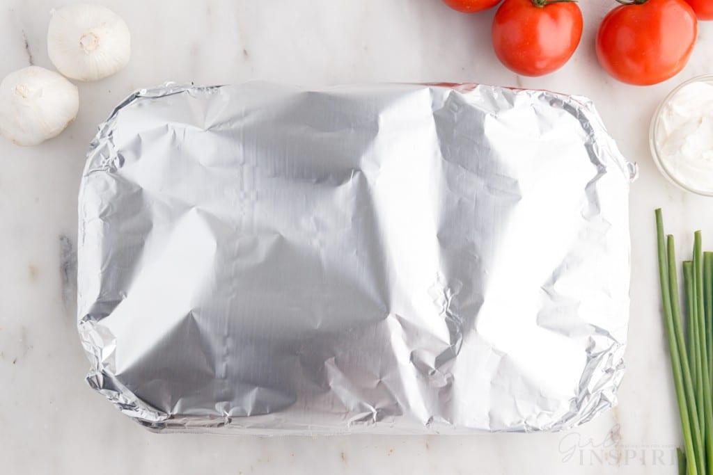 foil covering 9x13 next to sour cream, tomatoes, green onions, and garlic