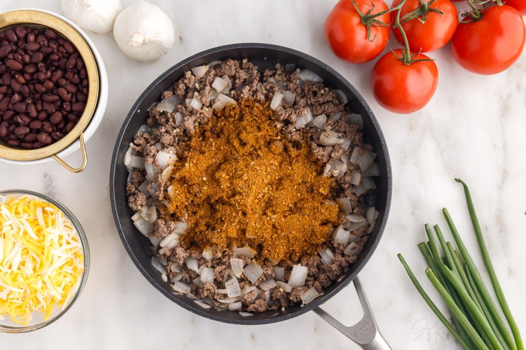 taco seasoning added to beef mixture in a skillet next to additional ingredients