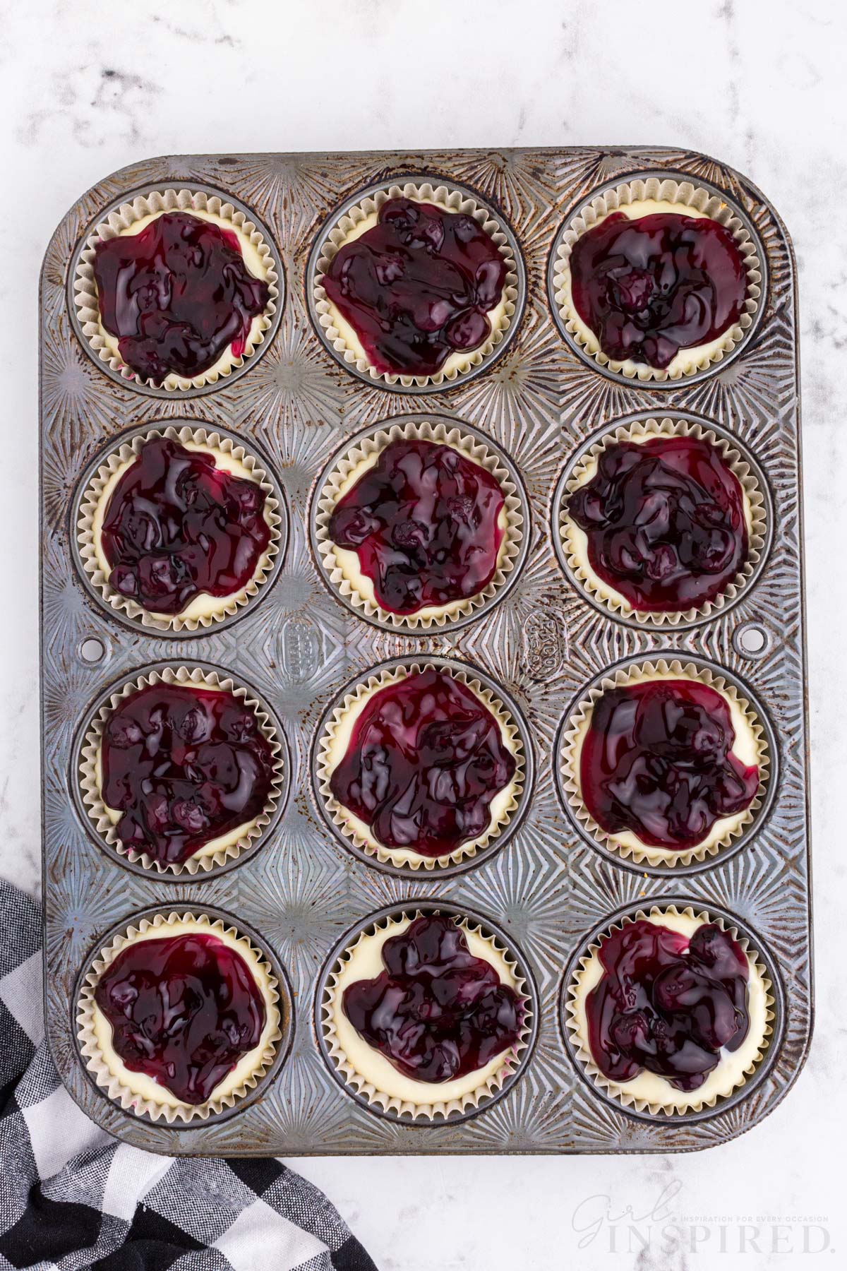 Metal muffin tray filled with Graham cracker crust, cheesecake filling and blueberry pie filling, checkered linen, on a marble countertop.