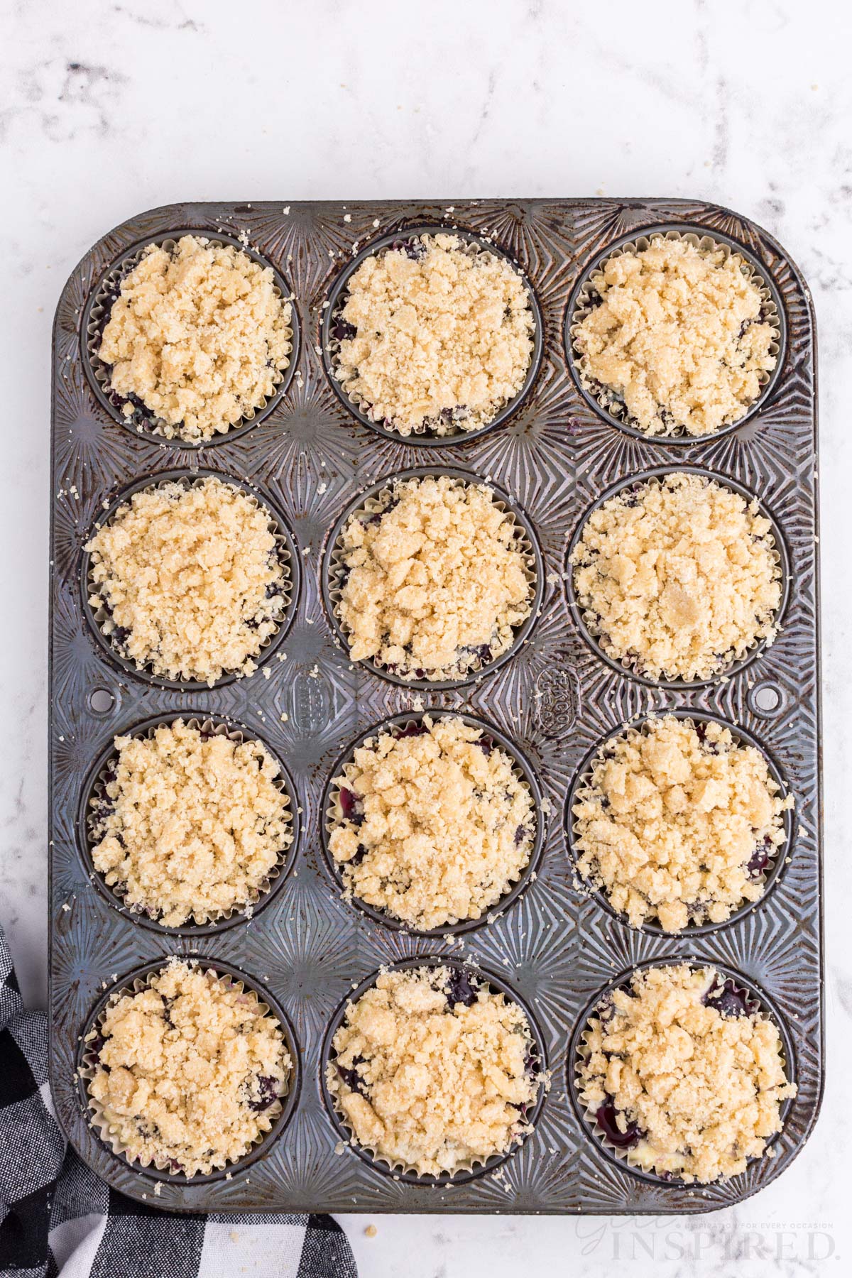 metal muffin tray with mini blueberry cheesecakes topped with streusel topping, checkered linen, on a marble countertop.