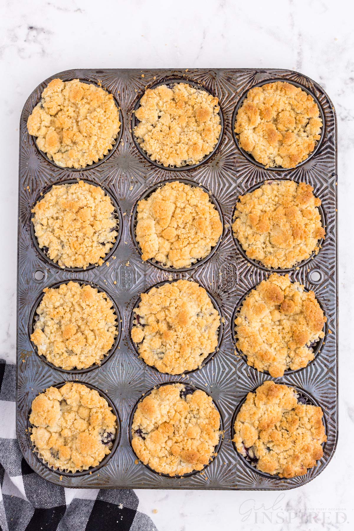 Metal muffin tray with baked mini blueberry cheesecakes, checkered linen, on a marble countertop.