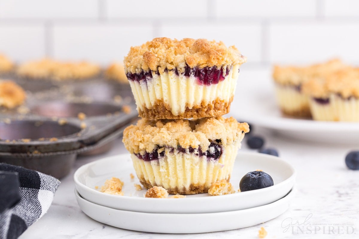 Two mini blueberry cheesecakes stacked on white serving plates, checkered linen, metal muffin tray with mini blueberry cheesecakes in the background, on a marble countertop.