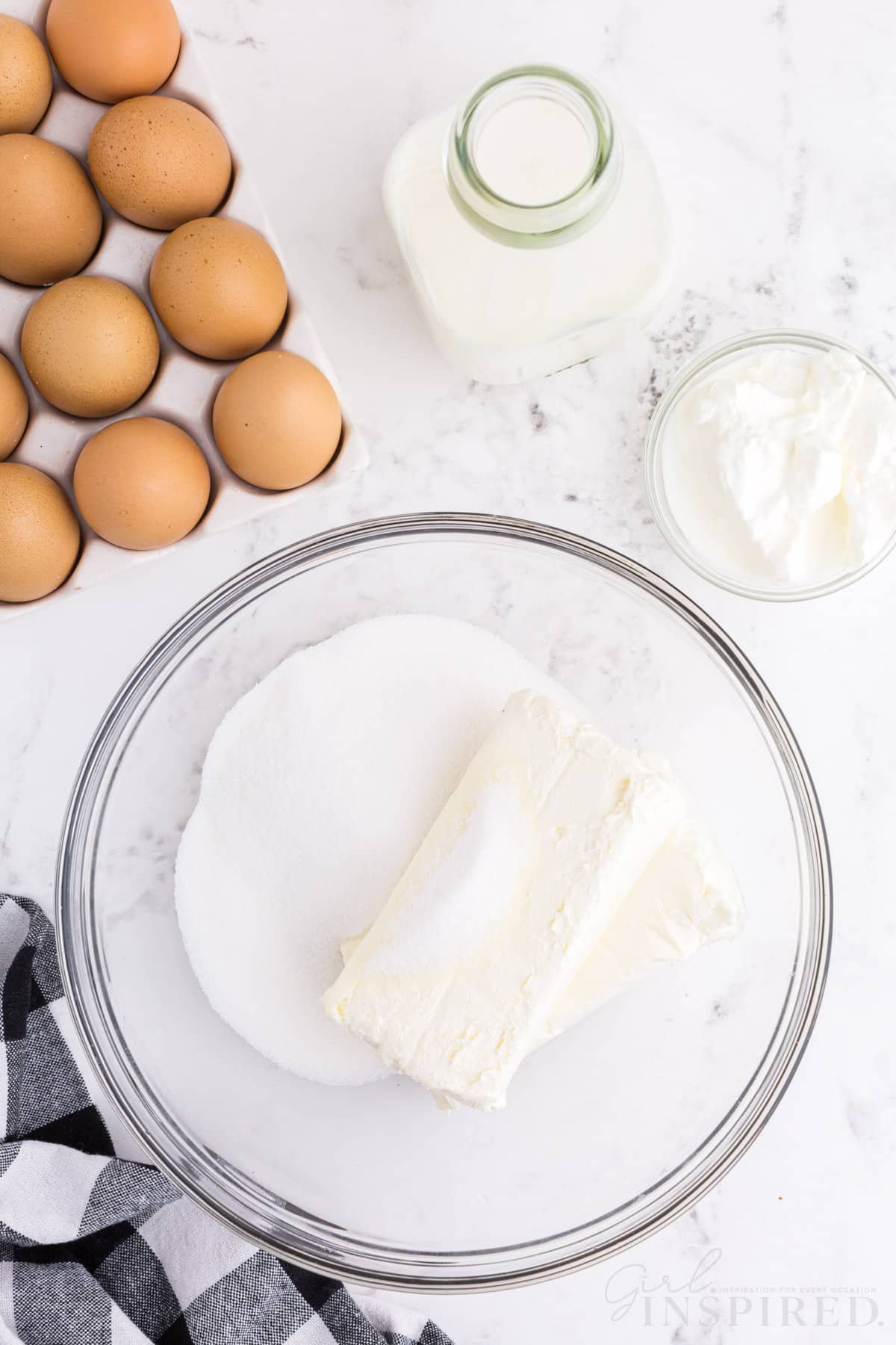 Mixing bowl with cream cheese and sugar, tray of eggs, checkered linen, jug of milk, small bowl of cream cheese, on a marble countertop.