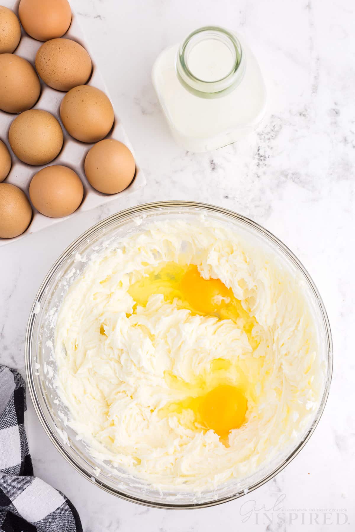 Bowl with cream cheese, sugar, and eggs, checkered linen, tray of eggs, jug of milk, on a marble countertop.