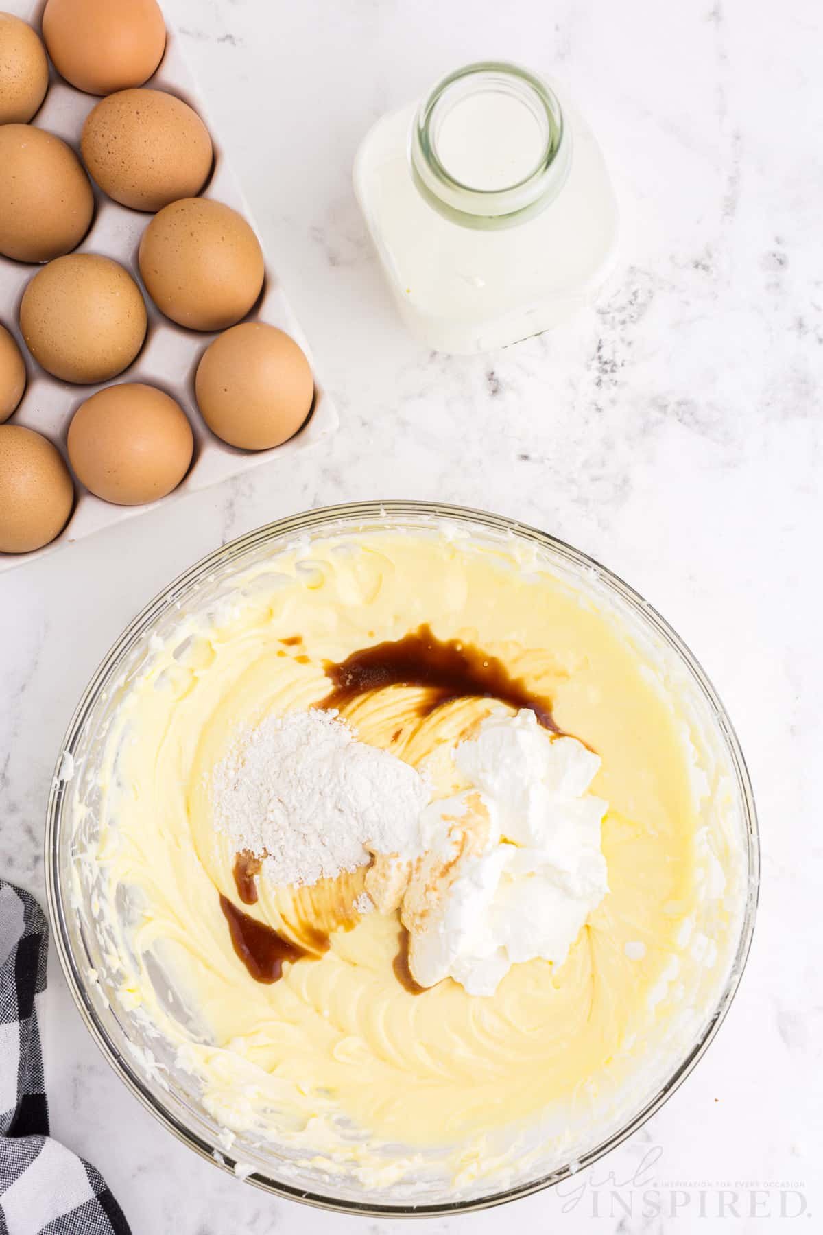 Mixing bowl with cheesecake mixture, sour cream, and vanilla extract, checkered linen, tray of eggs, jug of milk, on a marble countertop.