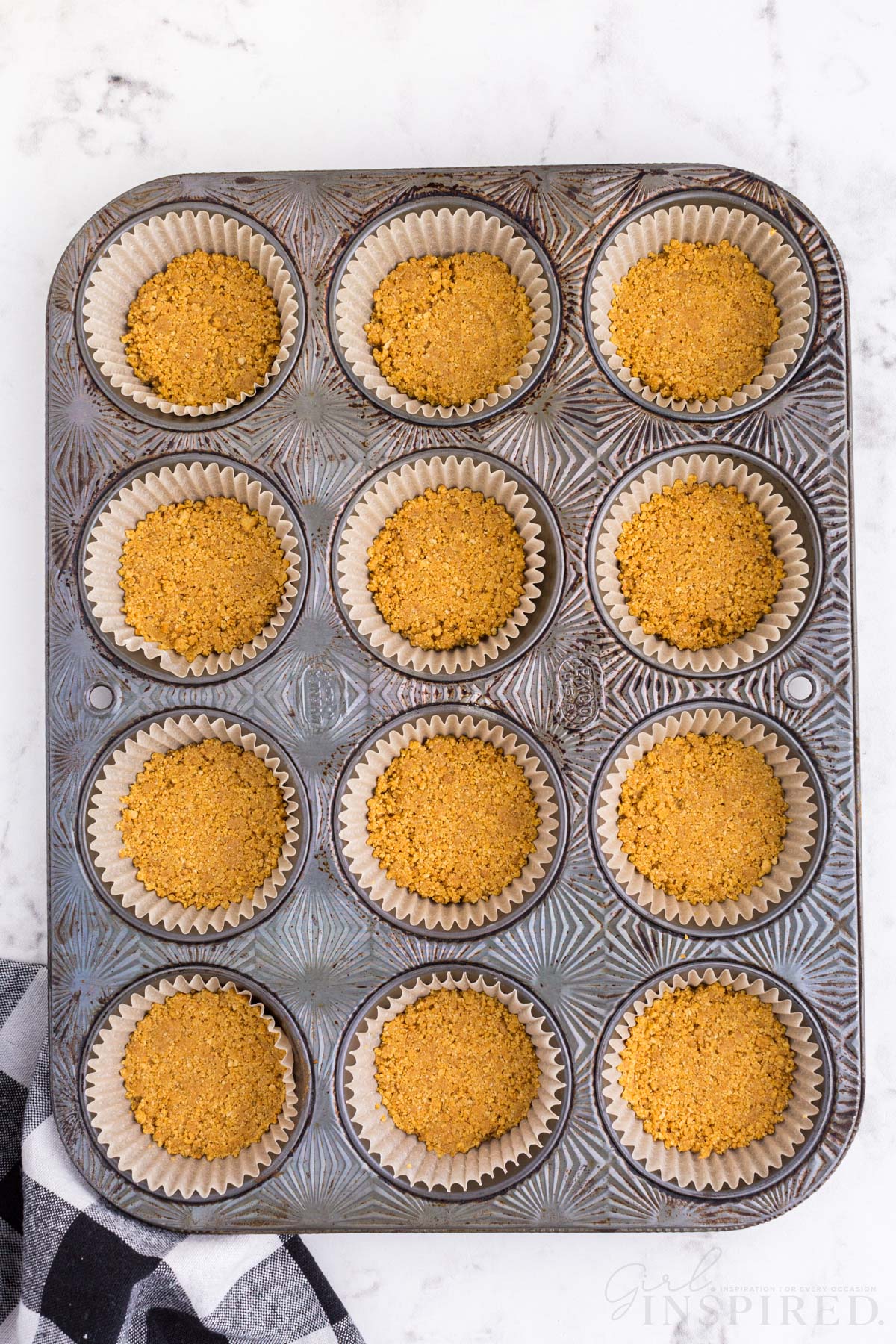 Metal cupcake tray with foil wrapper liners and Graham cracker crust pressed into each cup, checkered linen, on a marble countertop.
