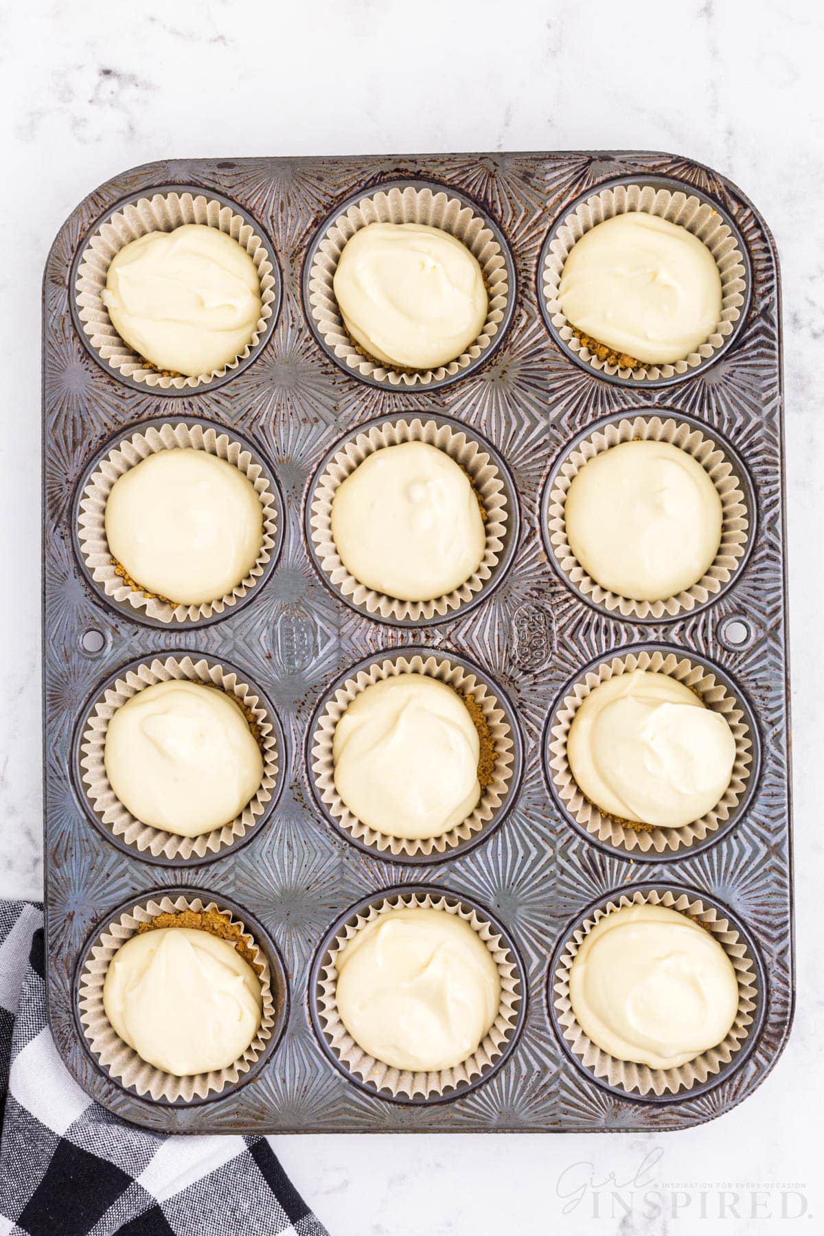 Metal muffin tray with cups filled with Graham cracker crust and cheesecake filling, checkered linen, on a marble countertop.