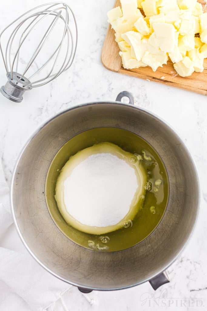 Metal mixing bowl with egg whites and sugar, wooden kitchen board with cubed butter, metal whisk attachment, on a marble countertop.