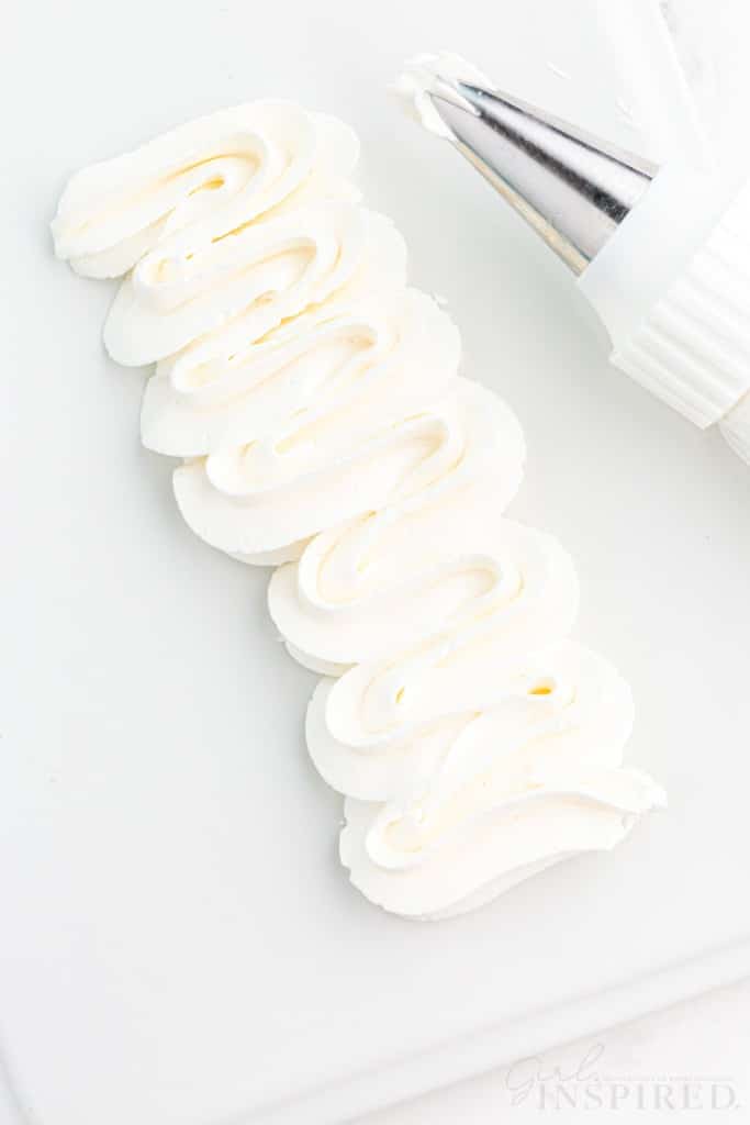 Close-up of piped Swiss meringue buttercream with piping nozzle on a white plastic board, on a marble countertop.