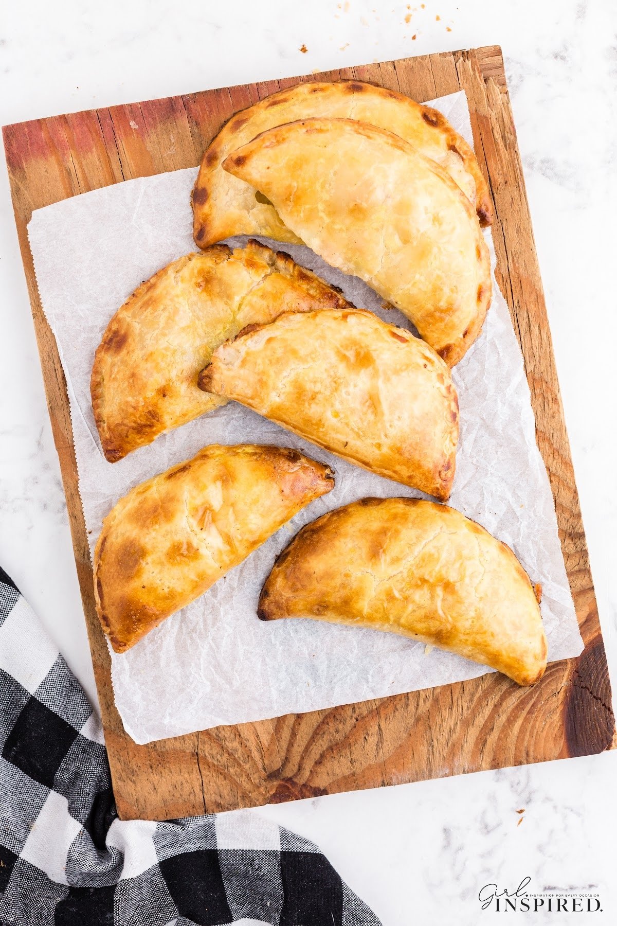 Six Chicken Pot Pie Hand Pies on a wooden kitchen board lined with parchment paper and a checkered cloth on the countertop.