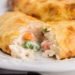 Chicken Pot Pie hand pie cut in half with filling coming out, atop a white serving plate with a tray of baked pies in the background.
