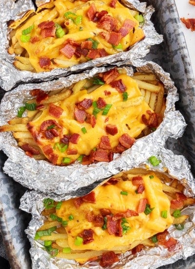Three foil packets with dirty fries in a baking tray, checkered linen, green onions, chopped bacon, on a marble countertop.