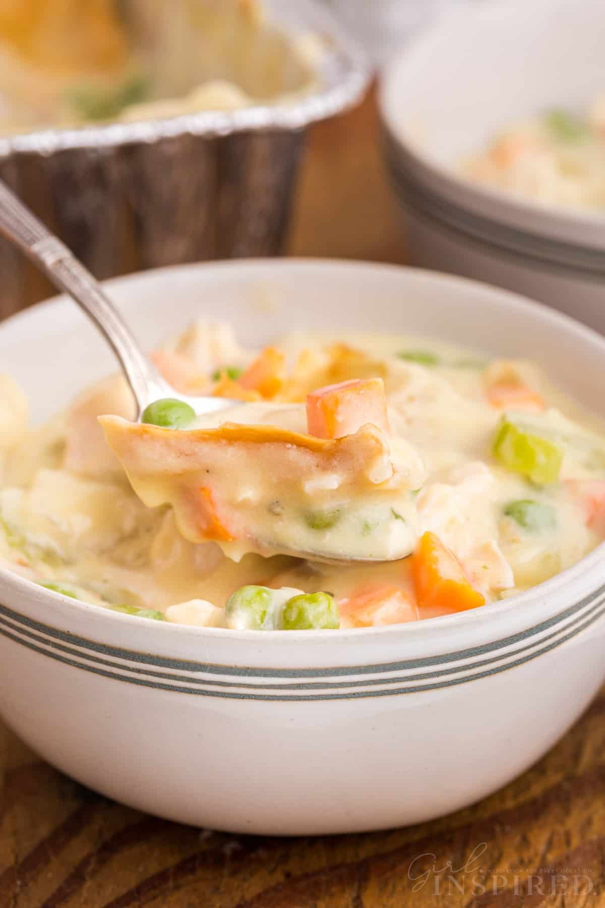 Spoon lifting a scoop of chicken pot pie from a bowl, freezer chicken pot pie in disposable container in background.