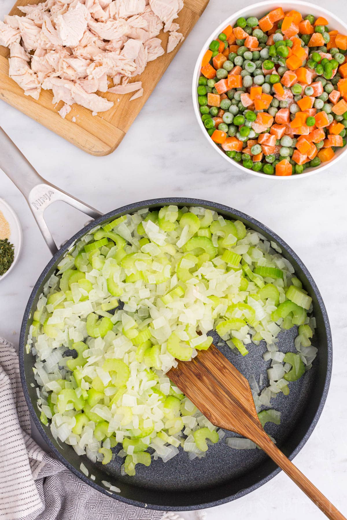 Heated skillet with melted butter, onions, and celery, diced cooked chicken on a wooden kitchen board, bowl of frozen peas and carrots.