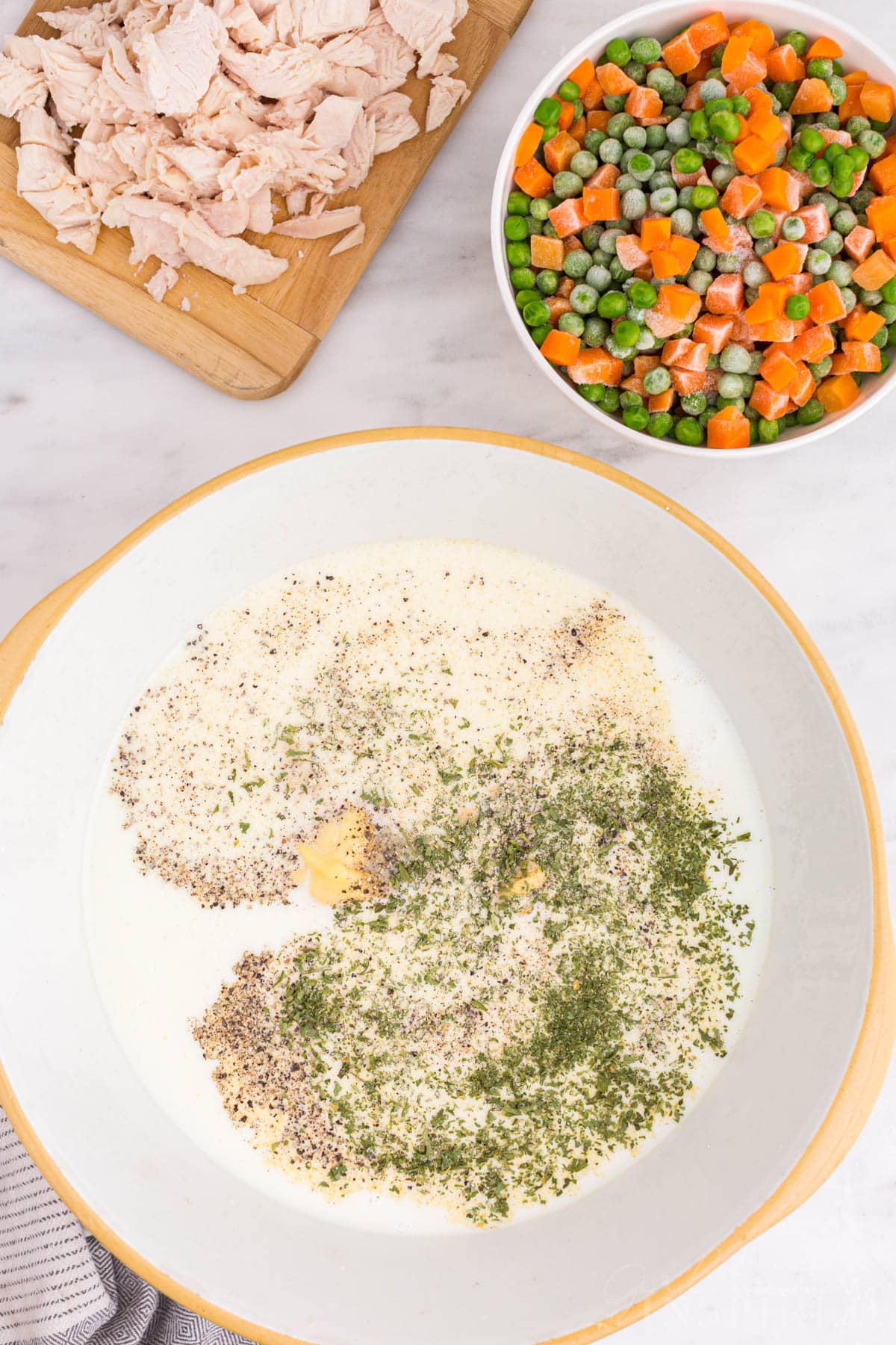 Bowl of milk, soup, and seasoning, bowl of frozen peas and carrots, diced chicken on a wooden kitchen board.