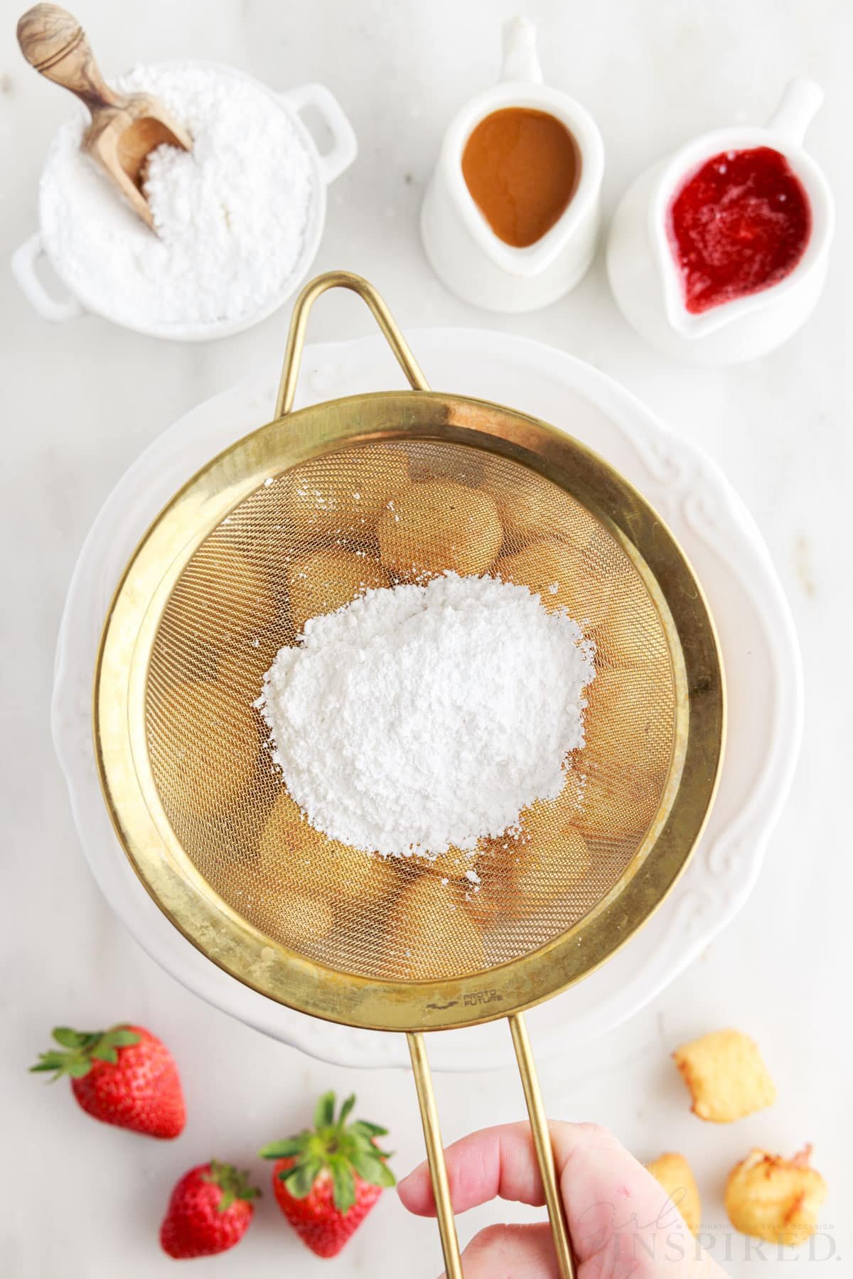 Powdered sugar in sifter over bowl of fried cheesecake bites.