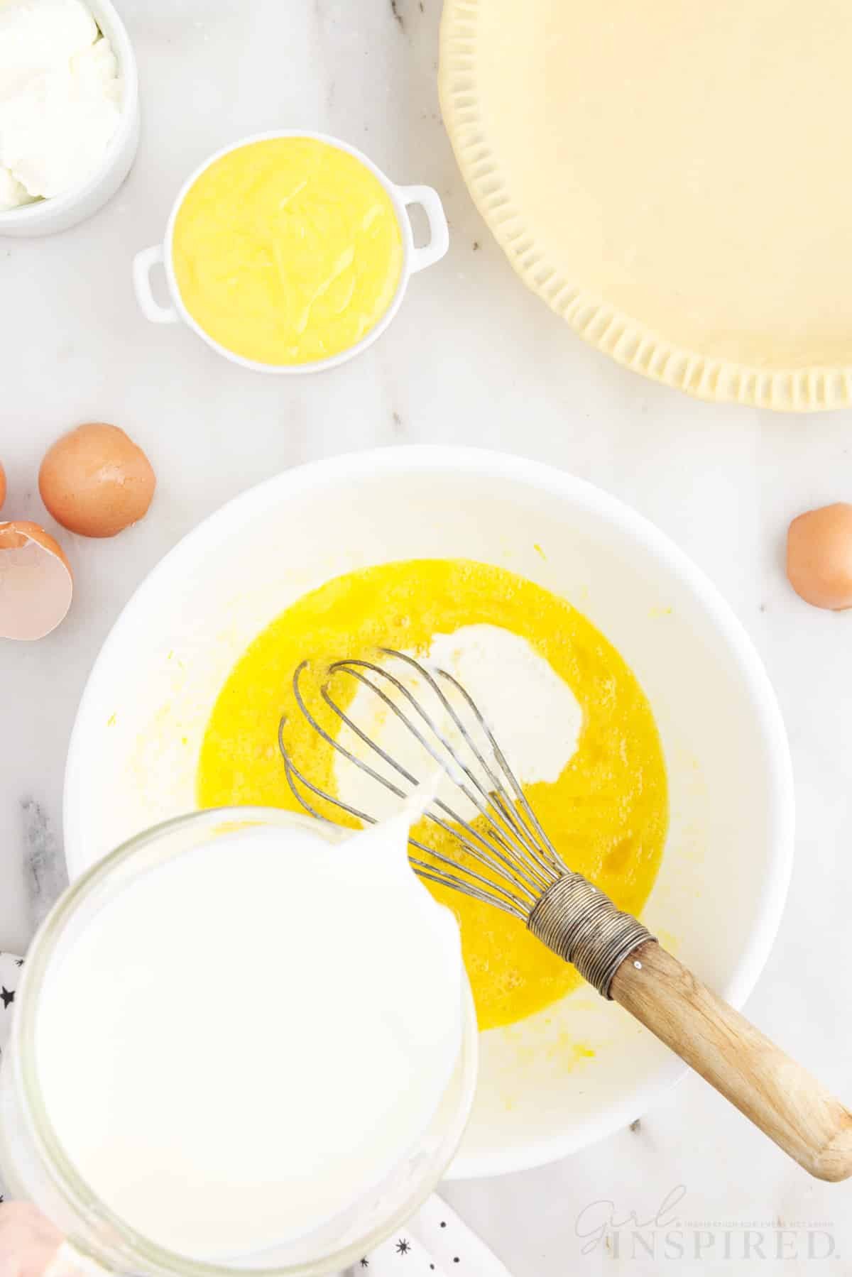 Milk added to egg mixture with a whisk inserted next to the pie crust for lemon custard pie.