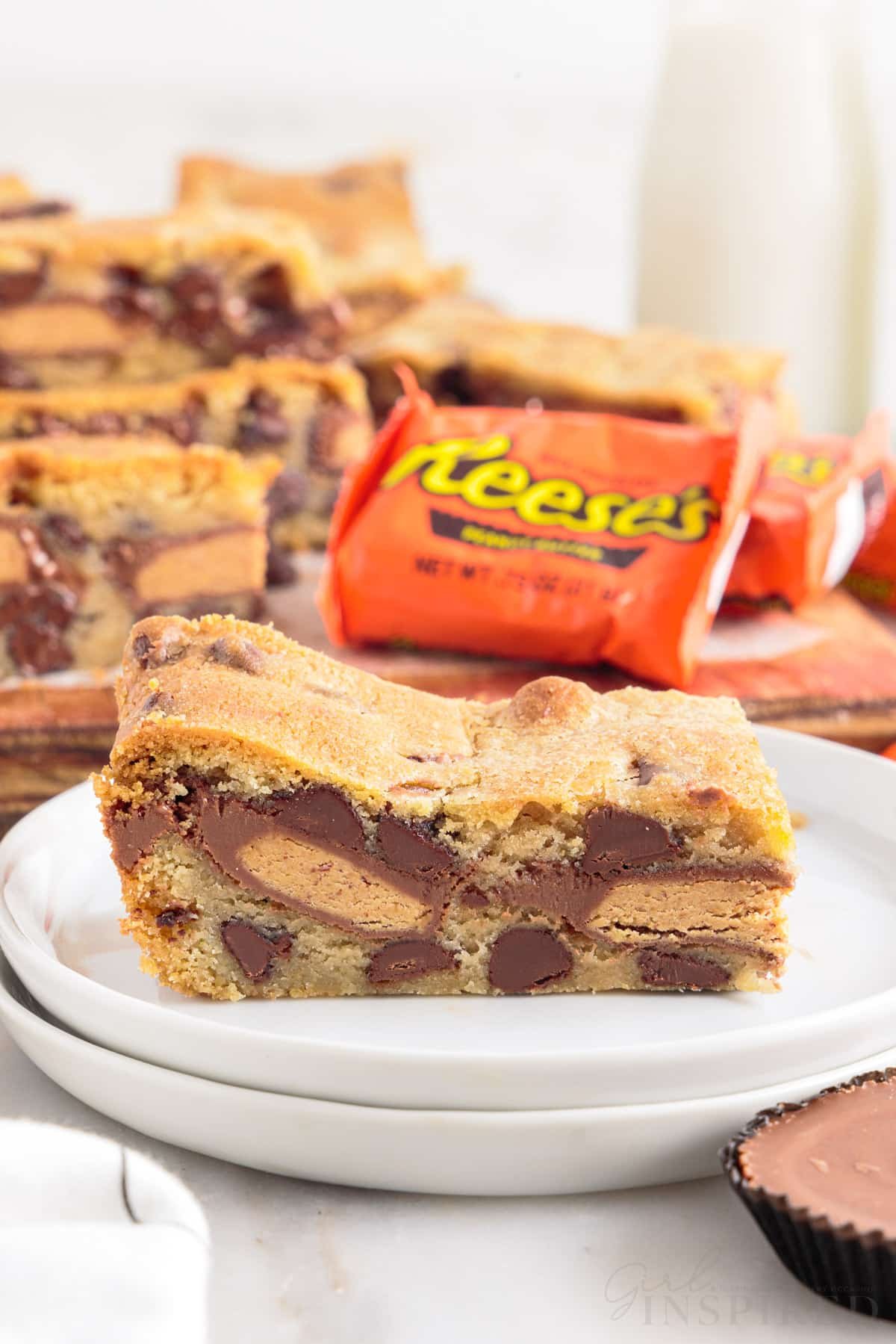 A small plate with a piece of a peanut butter cup cookie bar on it with Reese's in the background and additional bars.