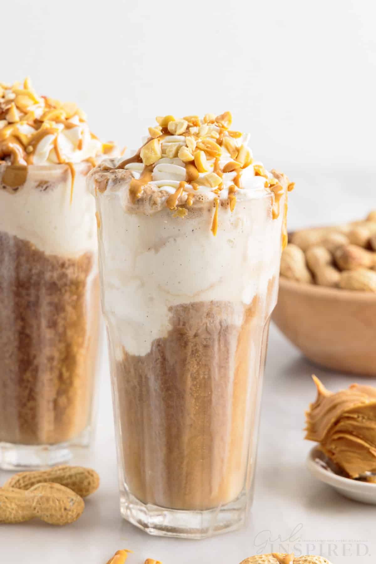 Two glasses of peanut butter root beer whiskey topped with whipped cream, peanuts, and peanut butter drizzle.