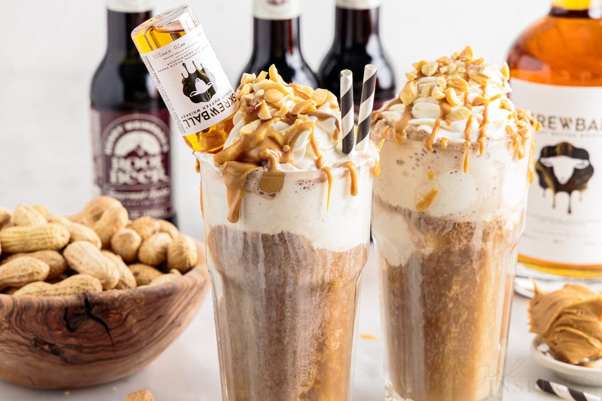 Two peanut butter root beer whiskeys with two straws and a mini bottle of Skrewball whiskey inserted in one next to a bowl of peanuts.