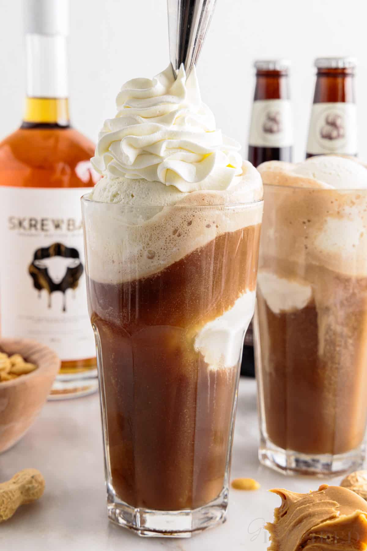 Whipped cream added to the top of a glass of peanut butter root beer whiskey with a second glass behind it.