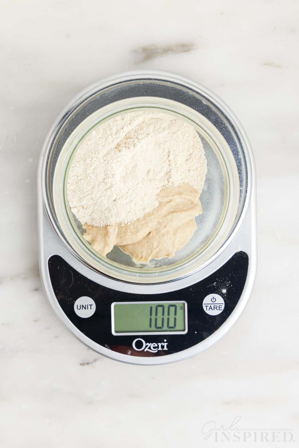 flour and sourdough starter mixture in a clean glass container on a scale that reads 100 grams.