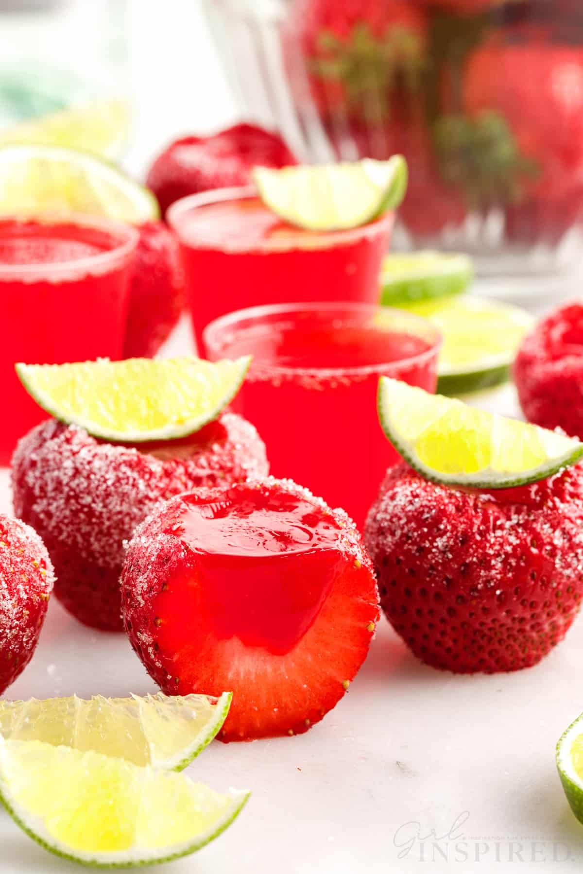 Strawberry daiquiri jello shots set on white counter with sugar coating and lime wedges as garnish; one sliced in half.