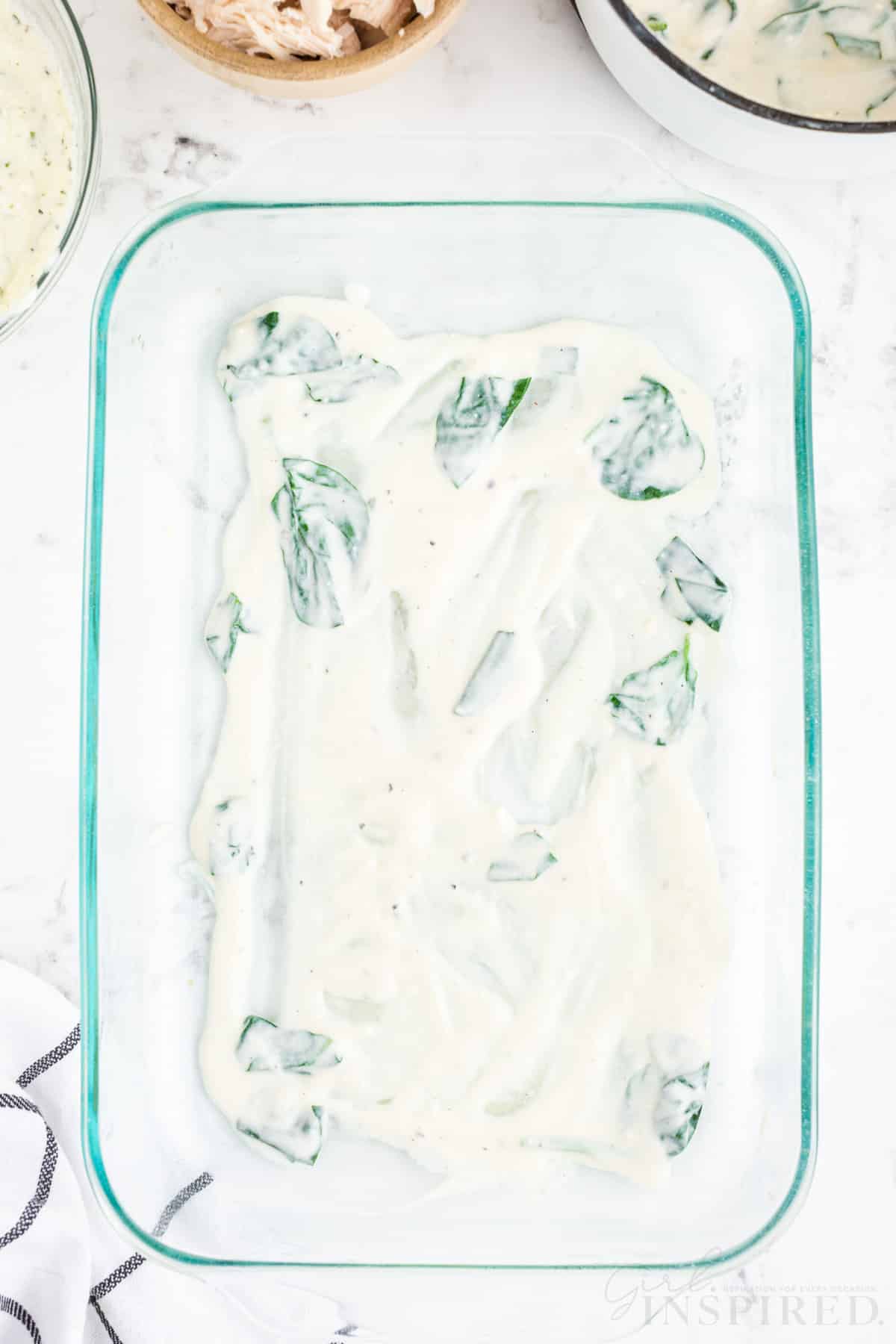 Layer of white lasagna sauce spread out in glass casserole dish, checkered linen, on a marble countertop.