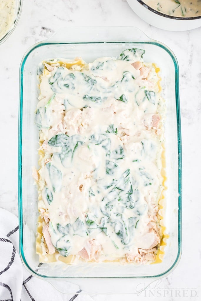Layer of white sauce on top of shredded chicken in glass casserole dish, checkered linen, on marble countertop.