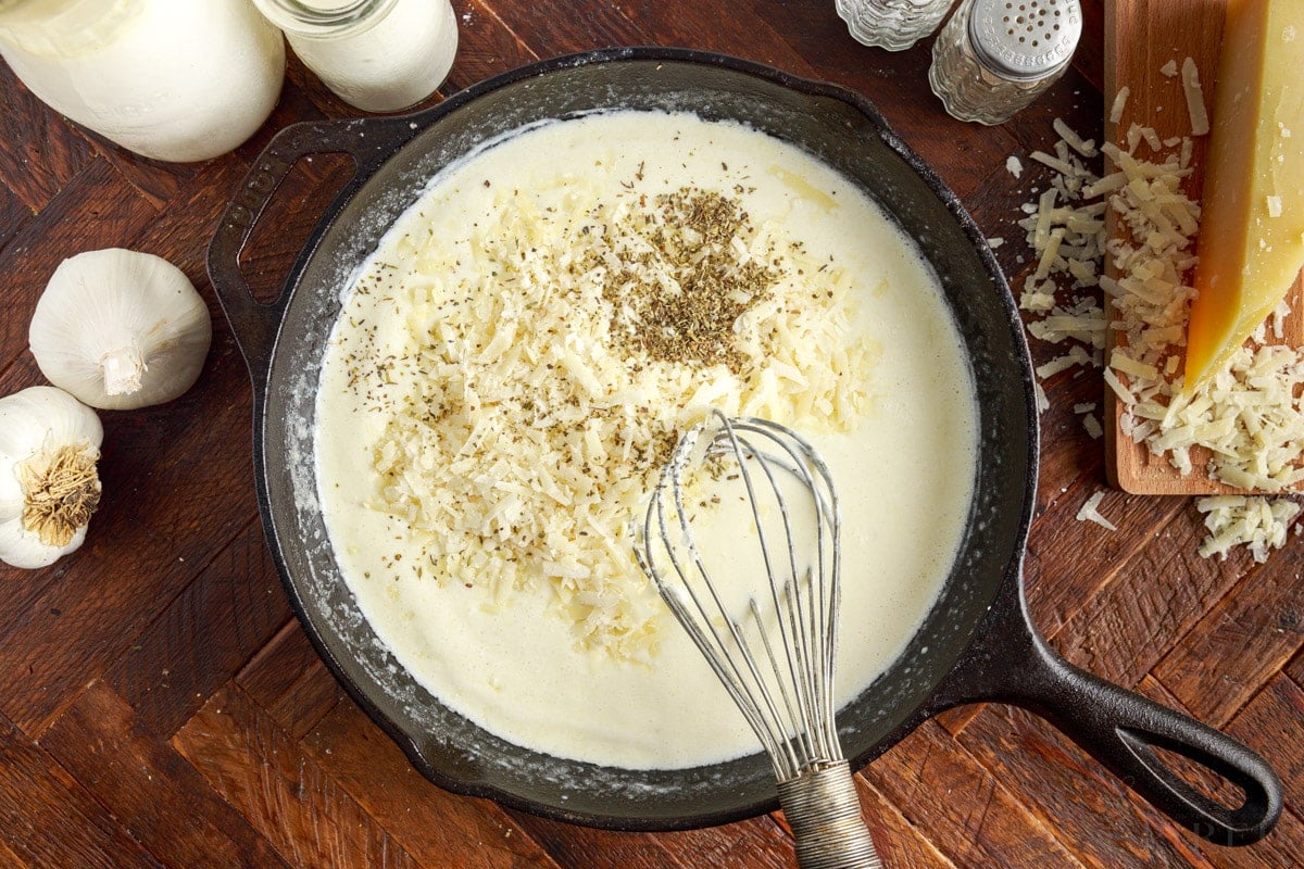 Grated parmesan cheese and dried herbs in skillet of fettuccine alfredo sauce with whisk.
