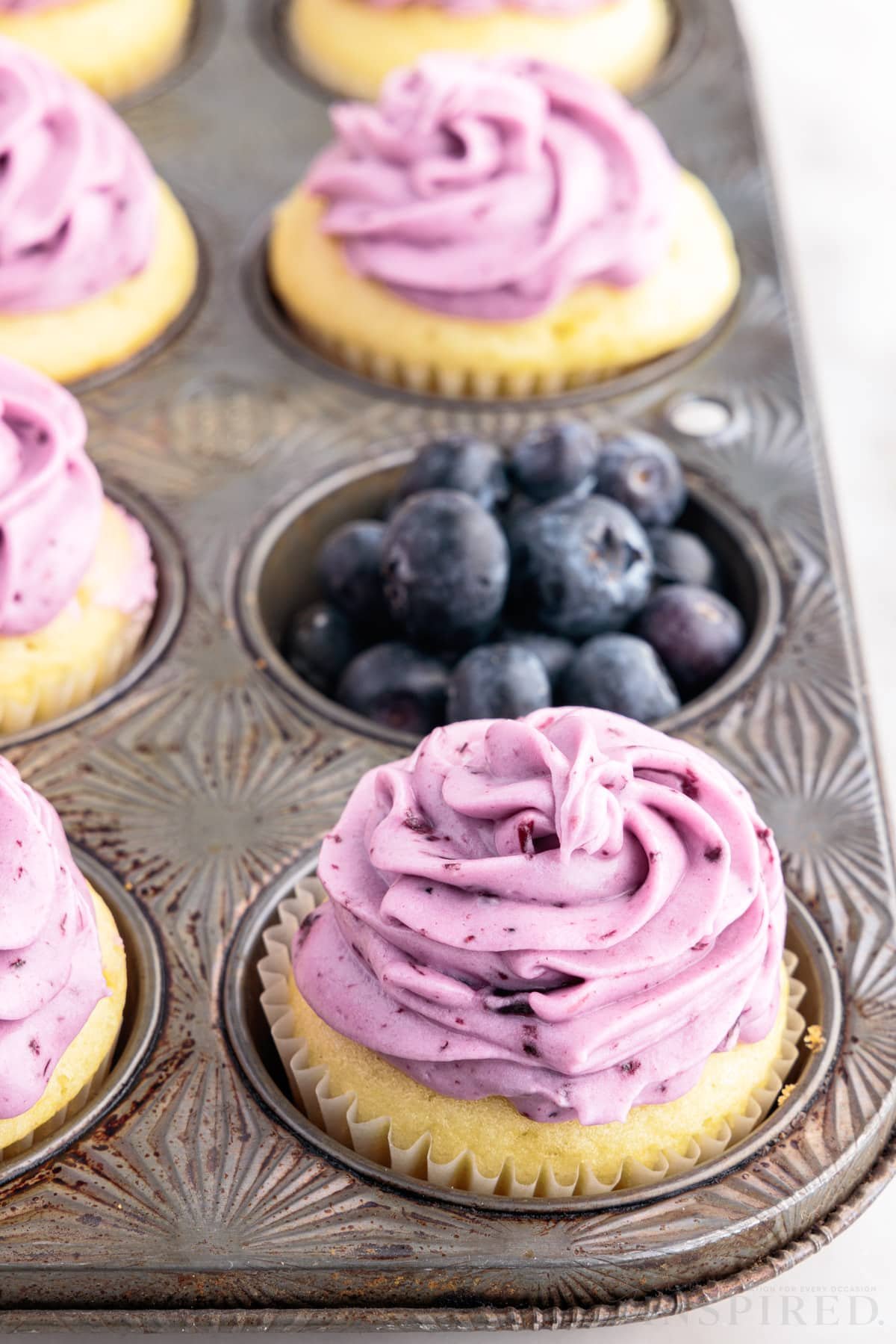 A muffin tin with Blueberry Cream Cheese Frosting covered cupcakes, one slot filled with blueberries.