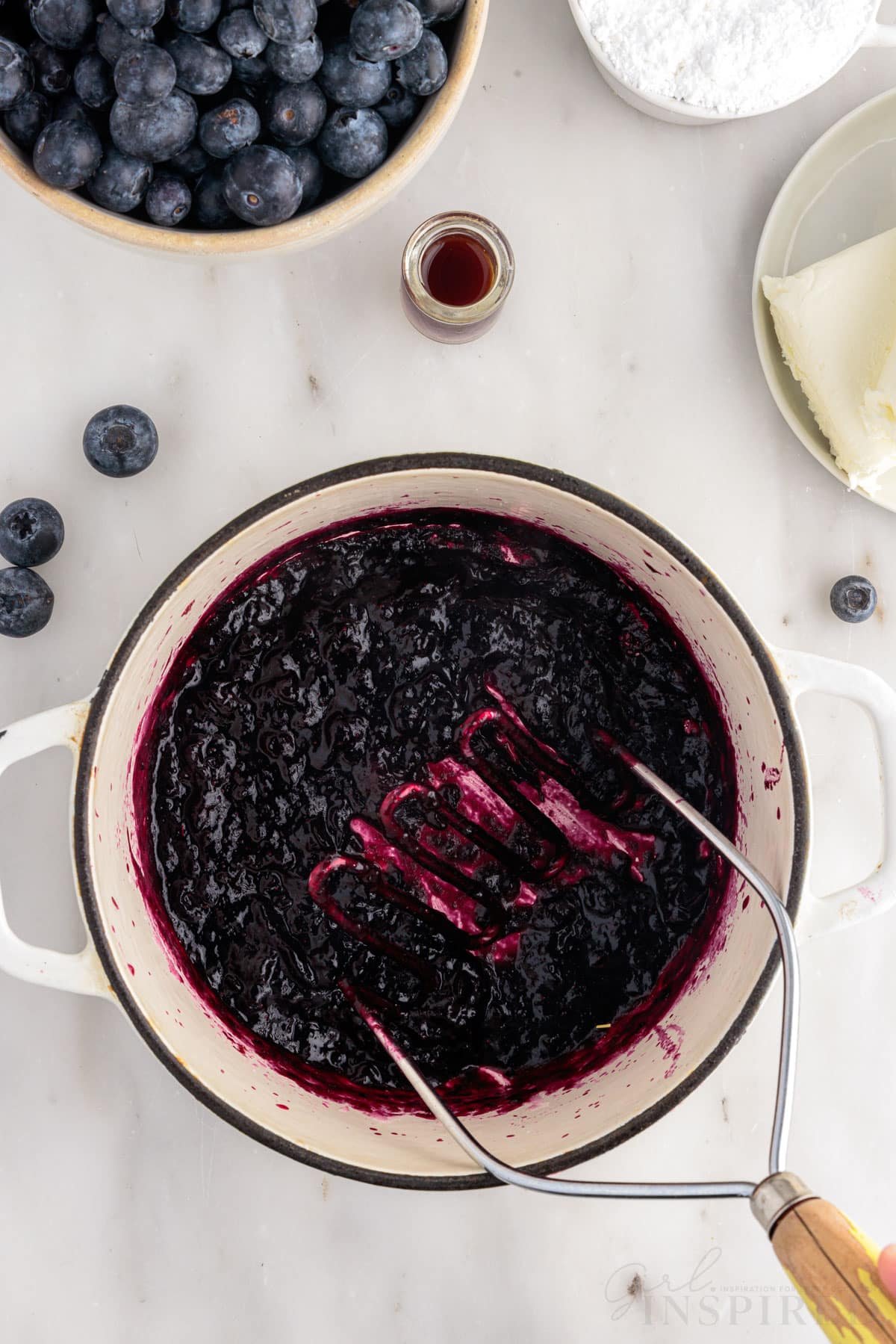 Dutch oven with blueberries being smashed with a potato masher to make Blueberry Cream Cheese Frosting.