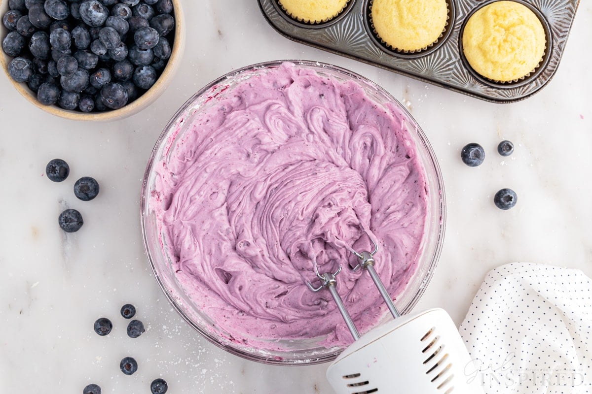 Overhead view of Blueberry Cream Cheese Frosting in a mixing bowl with a hand mixer next to a bowl of blueberries and a muffin tin full of cupcakes.