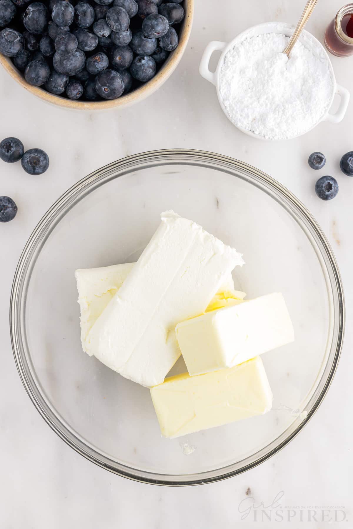 Cream cheese and butter in a mixing bowl next to powdered sugar and blueberries.