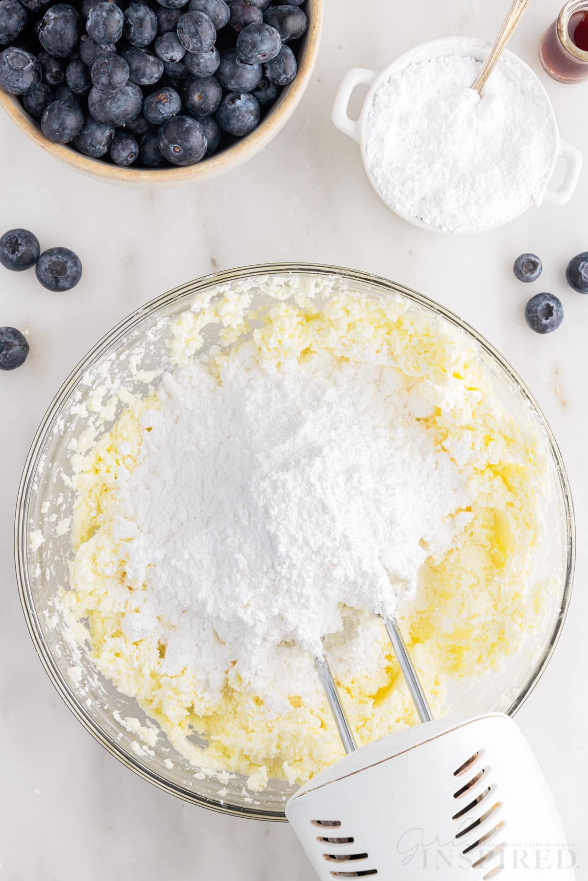 Powdered sugar added to butter mixture in a mixing bowl with a mixer inserted next to blueberries.