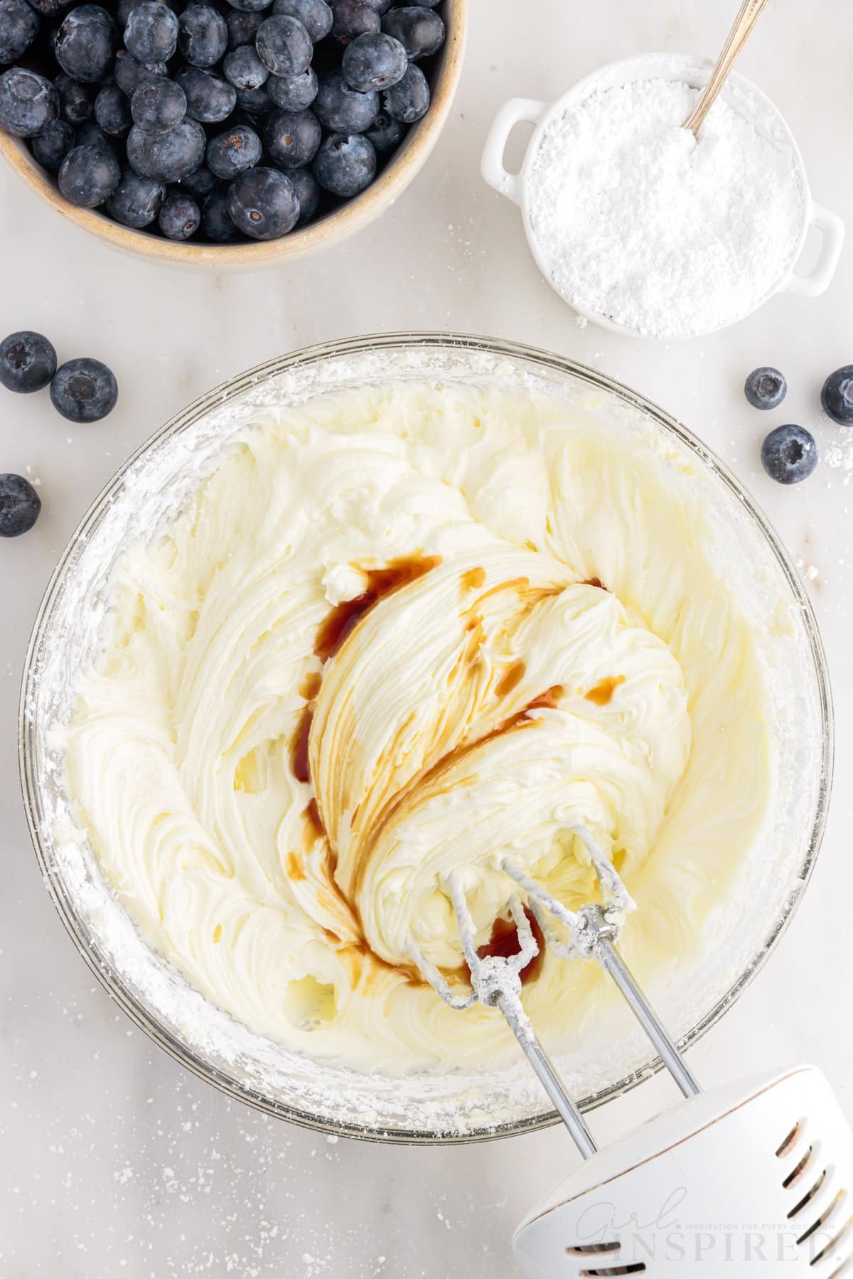 Vanilla added to frosting mixture with a hand mixer inserted next to blueberries in a bowl and a small dish of powdered sugar.