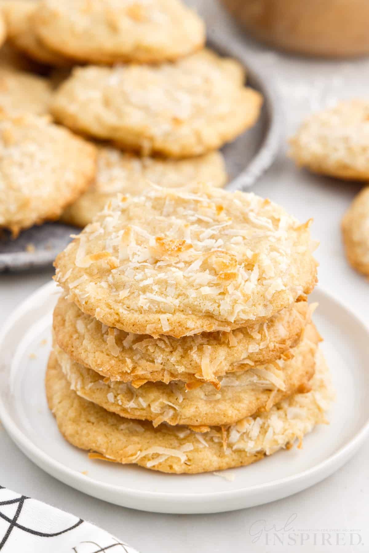 A small dish with four Chewy Coconut Cookies stacked on top of each other and a plate of them in the background.