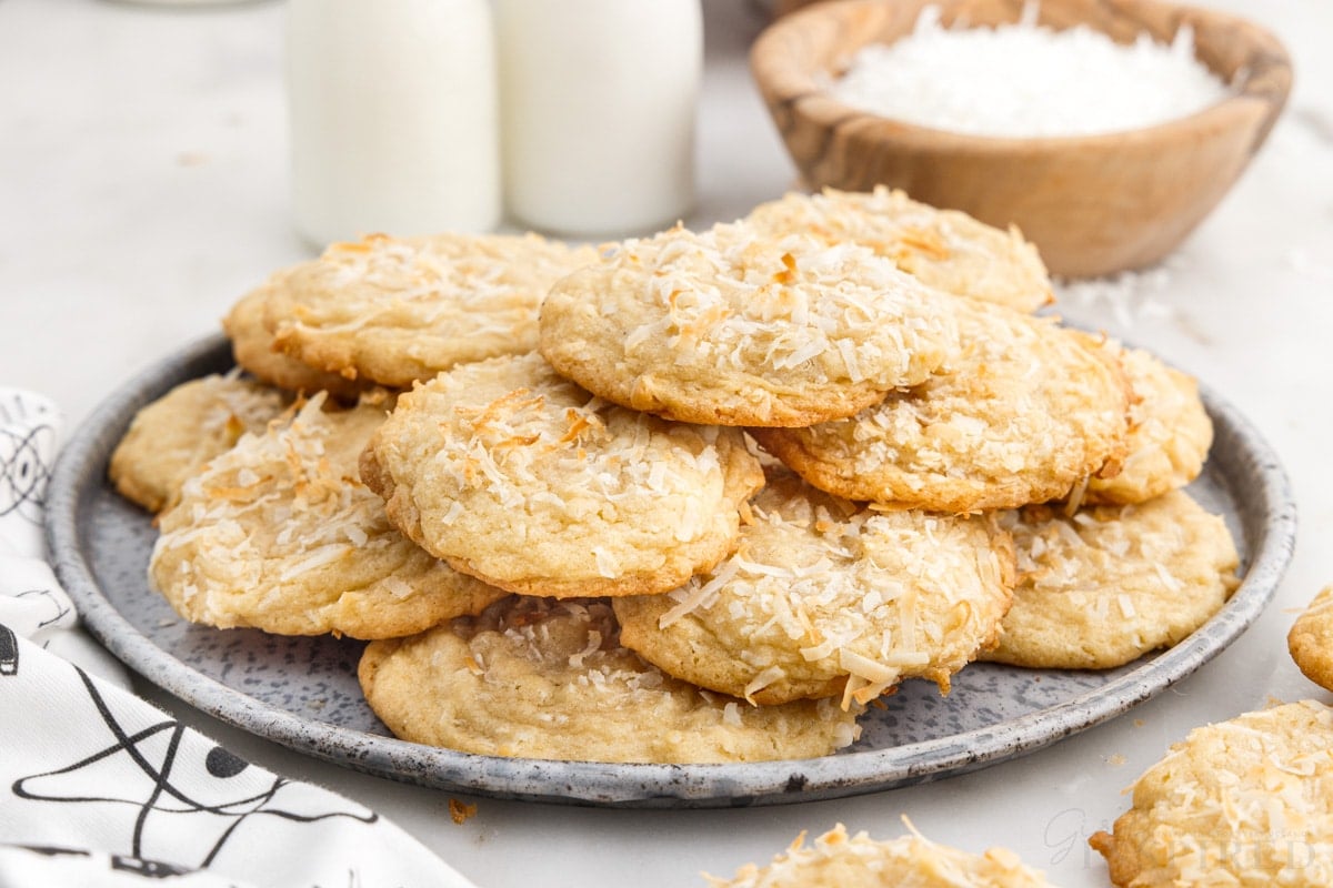 A plate of Chewy Coconut Cookies, shredded coconut in a bowl in the background next to two cups of milk.