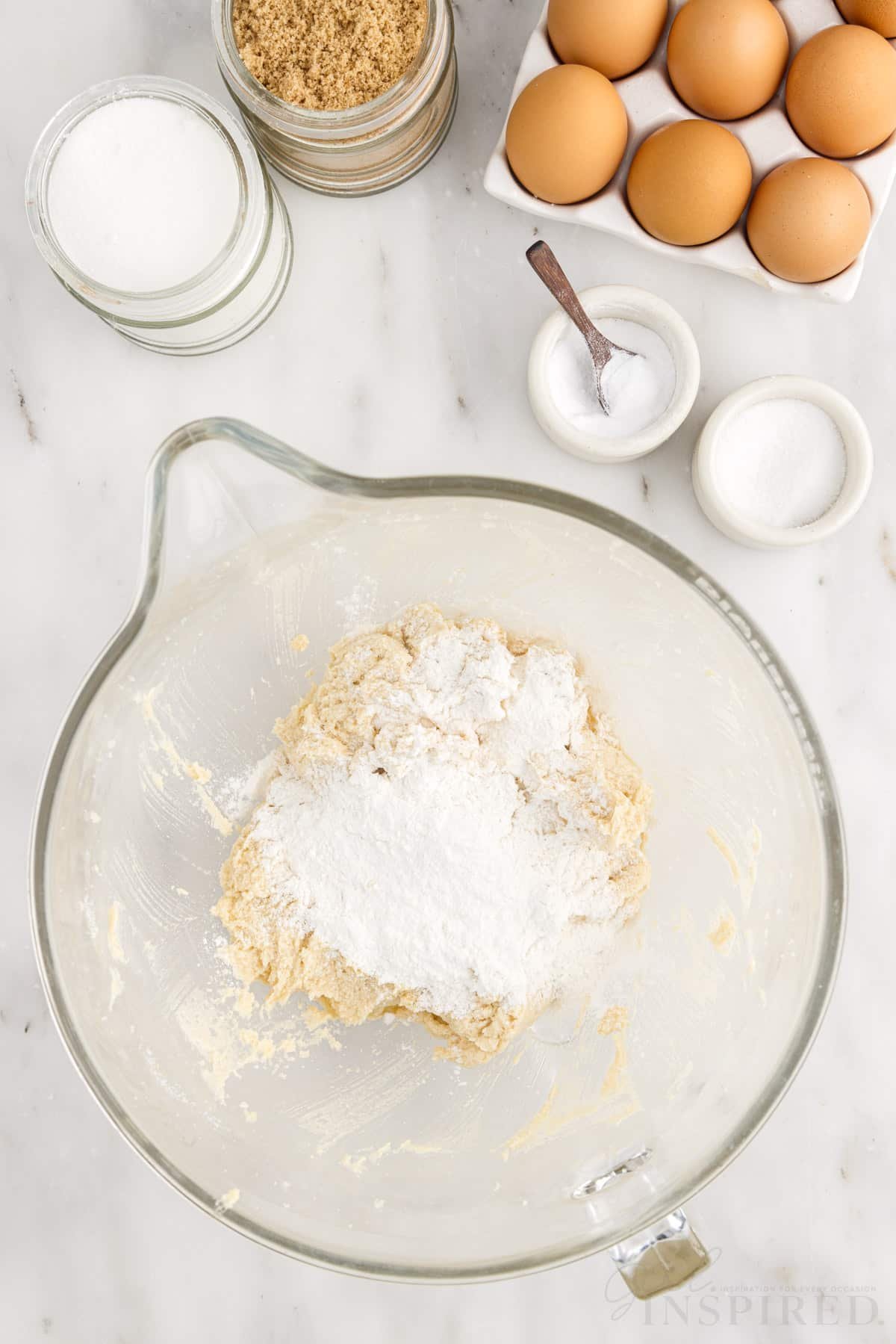 Flour mixture added to wet ingredients next to flour, eggs, and sugars.