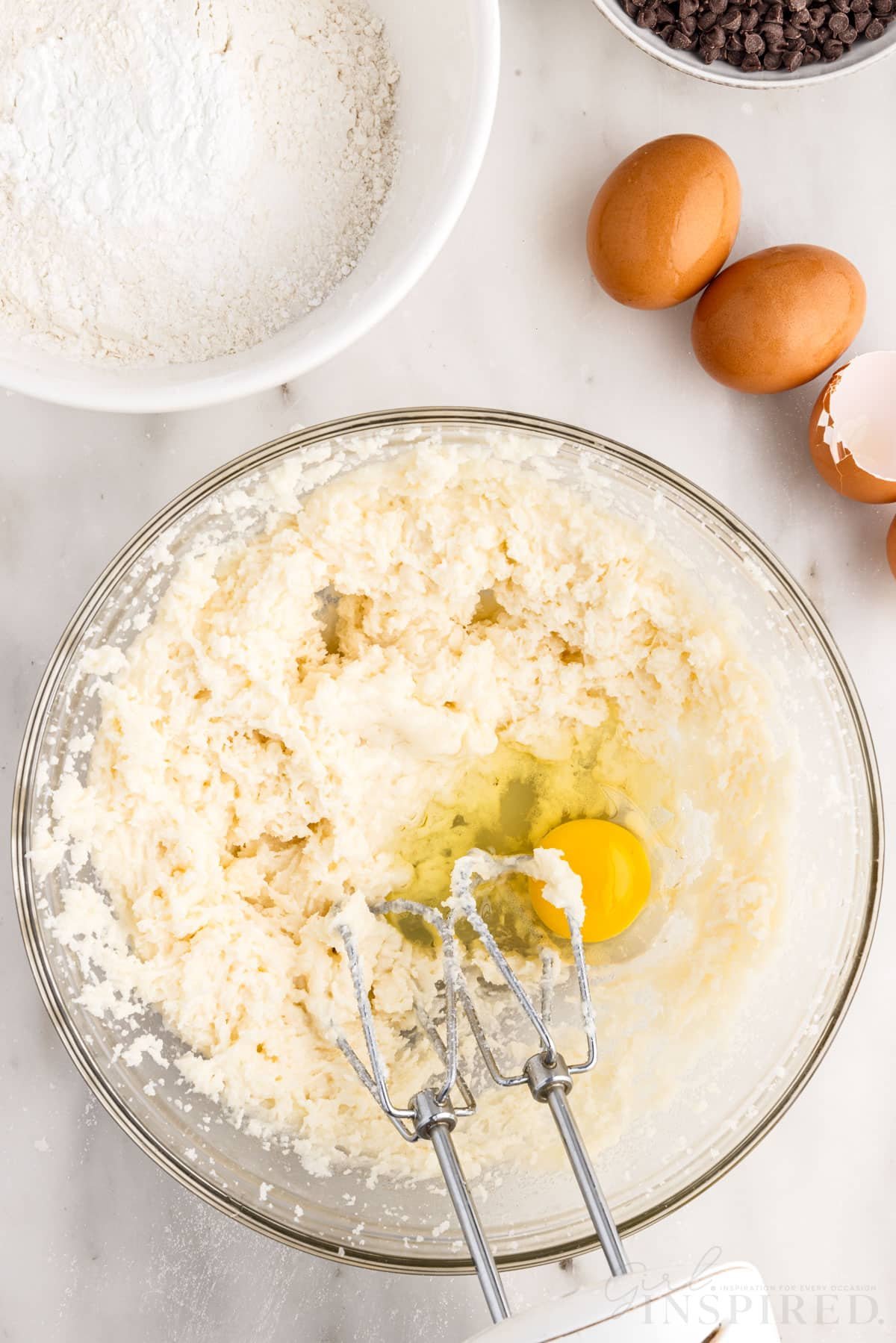 Egg added to batter next to eggs and bowl of flour with hand mixer inserted.