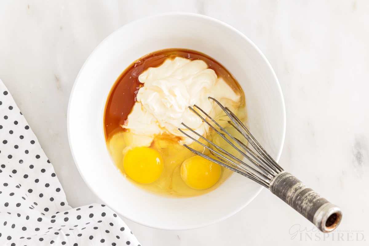 Mayonnaise, eggs, and vanilla added to milk in a mixing bowl with a whisk inserted.