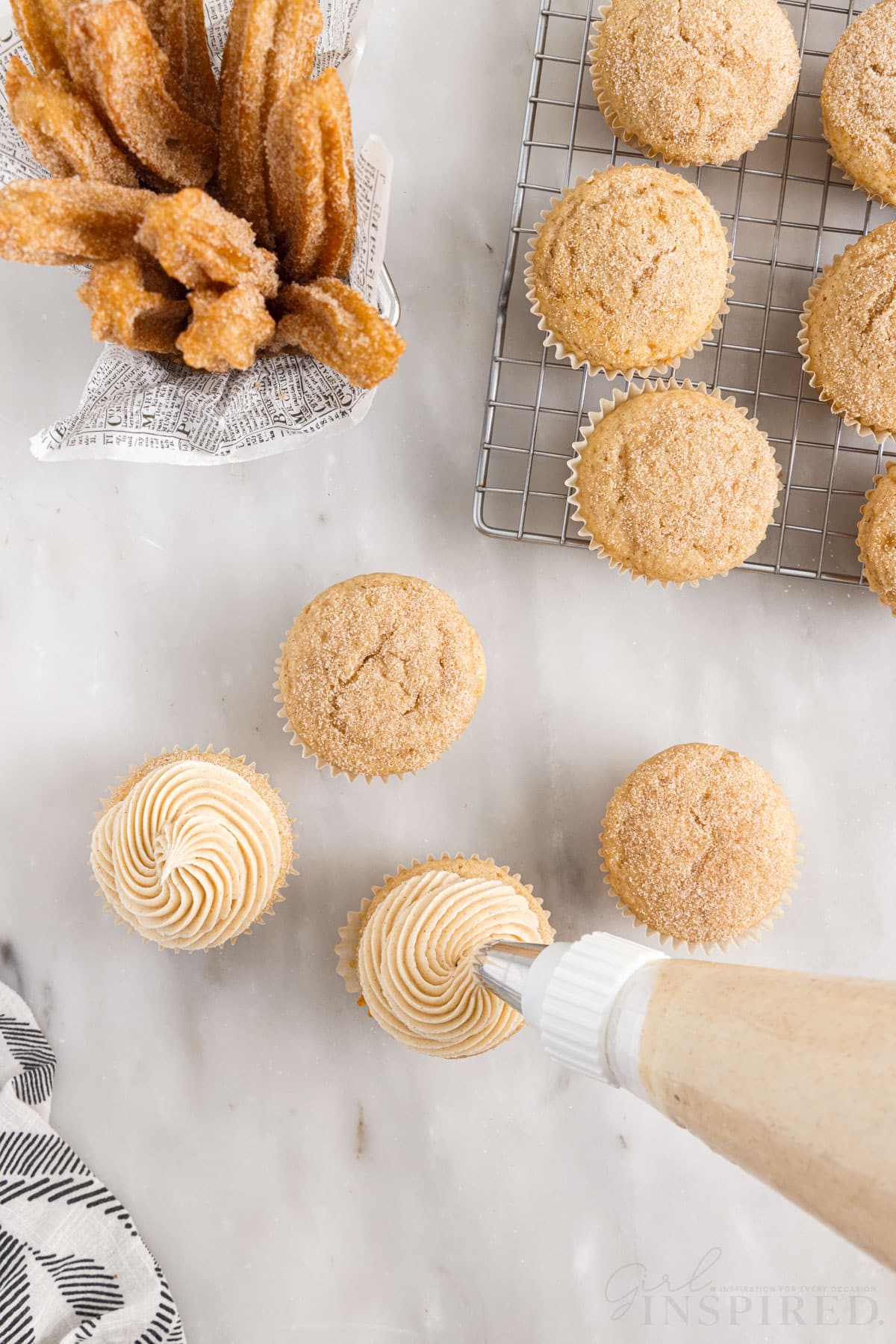 Four Churro Cupcakes being pipped with icing, in front of a cooling rack of Churro Cupcakes and a basket of Churros.