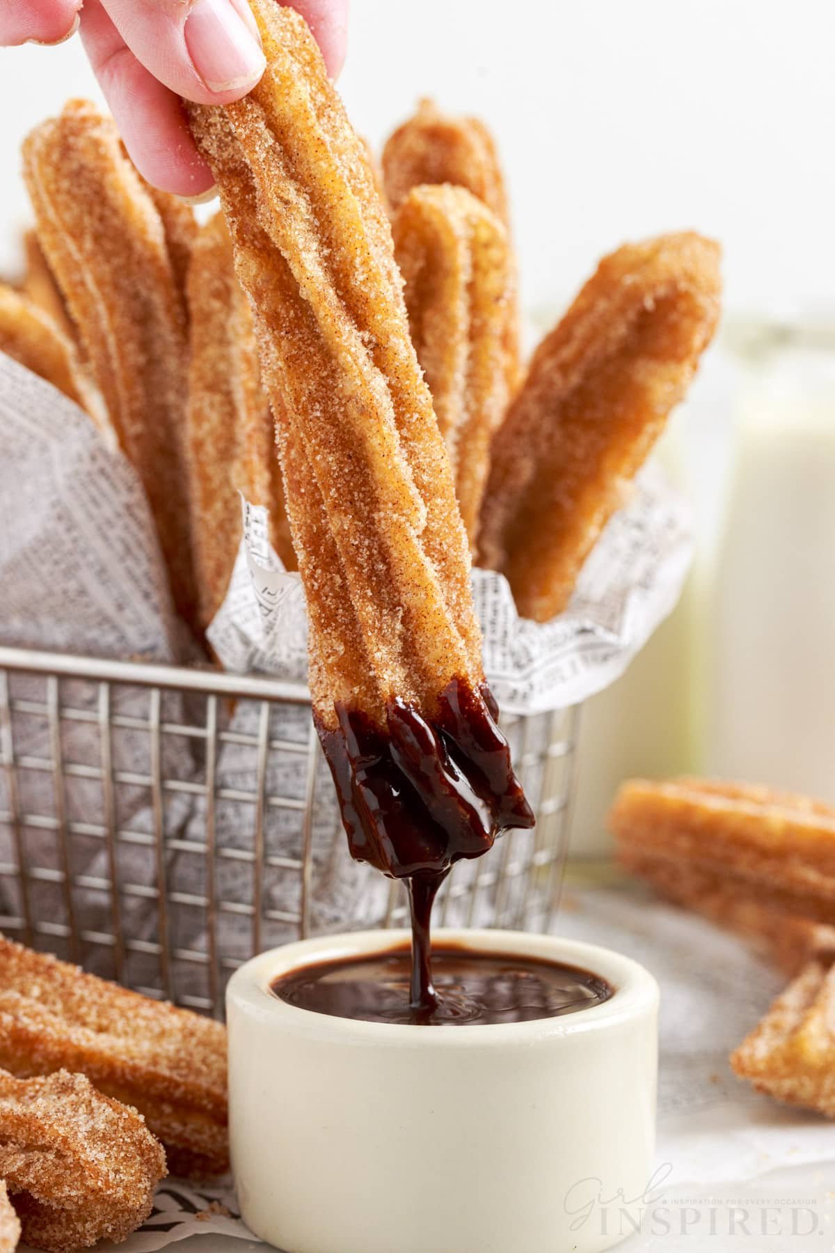 A Churro after being dipped in a small dish of chocolate, a wire basket in the background of Churros.